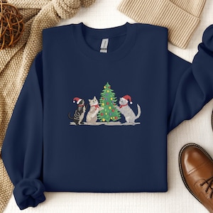 K&ouml;nnte beinhalten: Marineblaues Sweatshirt mit einer Stickerei von drei Katzen mit Weihnachtsm&uuml;tzen um einen geschm&uuml;ckten Weihnachtsbaum. Die Katzen sind grau, schwarz und wei&szlig;. Das Sweatshirt hat einen Rundhalsausschnitt und lange &Auml;rmel.
