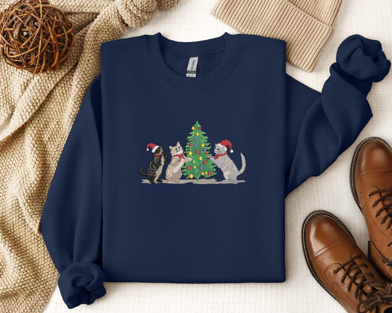 K&ouml;nnte beinhalten: Marineblaues Sweatshirt mit einer Stickerei von drei Katzen mit Weihnachtsm&uuml;tzen um einen geschm&uuml;ckten Weihnachtsbaum. Die Katzen sind grau, schwarz und wei&szlig;. Das Sweatshirt hat einen Rundhalsausschnitt und lange &Auml;rmel.