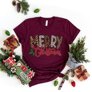 May include: A maroon t-shirt with the words "Merry Christmas" in a leopard print design. The words are in a red and white font with a green holly leaf and red berries.