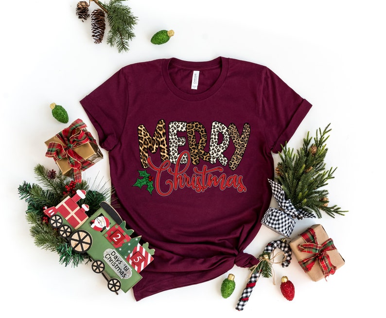 May include: A maroon t-shirt with the words "Merry Christmas" in a leopard print design. The words are in a red and white font with a green holly leaf and red berries.