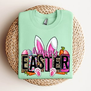 May include: A light green sweatshirt with a graphic design of a bunny with pink ears and the words "Happy Easter" in black letters. The graphic also includes colorful eggs, carrots, and a leopard print pattern.