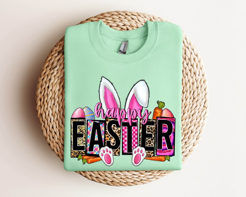 May include: A light green sweatshirt with a graphic design of a bunny with pink ears and the words "Happy Easter" in black letters. The graphic also includes colorful eggs, carrots, and a leopard print pattern.