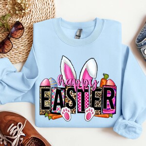 May include: Light blue sweatshirt with a graphic design of a bunny with pink ears and the text "Happy Easter" in black letters. The graphic also includes colorful eggs, carrots, and a checkered pattern.