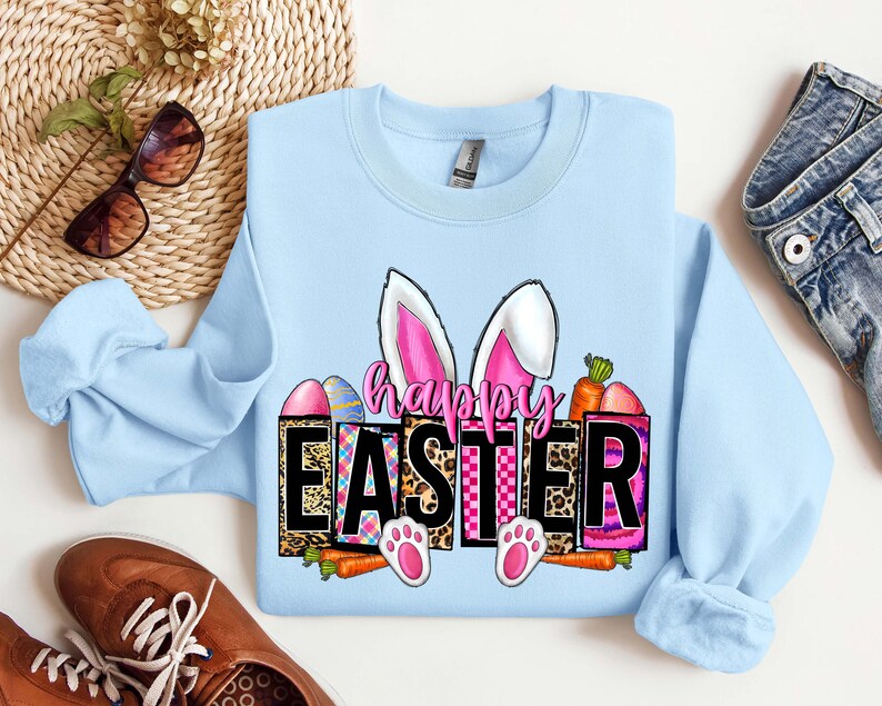 May include: Light blue sweatshirt with a graphic design of a bunny with pink ears and the text "Happy Easter" in black letters. The graphic also includes colorful eggs, carrots, and a checkered pattern.
