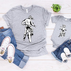 May include: Two gray t-shirts with a graphic of a person riding a dirt bike. The larger shirt has a graphic of a person riding a dirt bike with the text "Dirt Bike Dad". The smaller shirt has a graphic of a person riding a dirt bike with the text "Dirt Bike Kid".