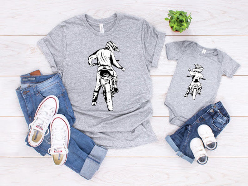 May include: Two gray t-shirts with a graphic of a person riding a dirt bike. The larger shirt has a graphic of a person riding a dirt bike with the text "Dirt Bike Dad". The smaller shirt has a graphic of a person riding a dirt bike with the text "Dirt Bike Kid".