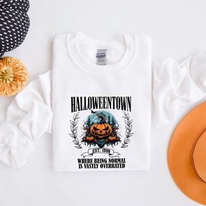 Może przedstawiać: Biała bluza z nadrukiem graficznym przedstawiającym dynię z przerażającą twarzą i tekstem "HALLOWEENTOWN EST. 1998 WHERE BEING NORMAL IS VASTLY OVERRATED".