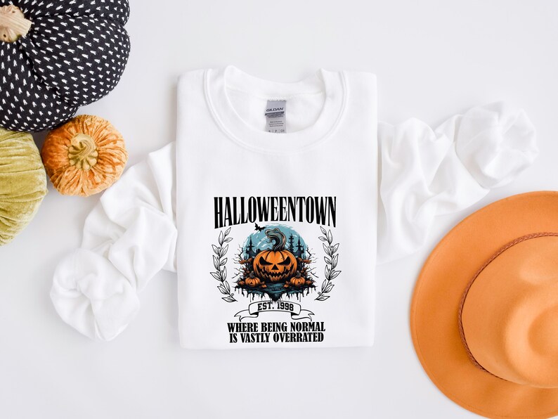 Może przedstawiać: Biała bluza z nadrukiem graficznym przedstawiającym dynię z przerażającą twarzą i tekstem "HALLOWEENTOWN EST. 1998 WHERE BEING NORMAL IS VASTLY OVERRATED".