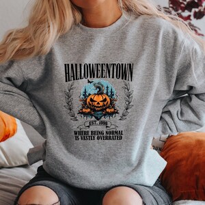 Może przedstawiać: Szara bluza z nadrukiem graficznym przedstawiającym dynię i tekstem "Halloweentown Est. 1998 Where being normal is vastly overrated".