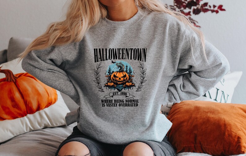 Może przedstawiać: Szara bluza z nadrukiem graficznym przedstawiającym dynię i tekstem "Halloweentown Est. 1998 Where being normal is vastly overrated".