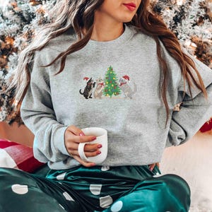 K&ouml;nnte beinhalten: Ein graues Sweatshirt mit einem gestickten Design von drei Katzen mit Weihnachtsm&uuml;tzen um einen Weihnachtsbaum. Die Katzen stehen vor einem geschm&uuml;ckten Weihnachtsbaum. Die Person h&auml;lt eine wei&szlig;e Tasse.