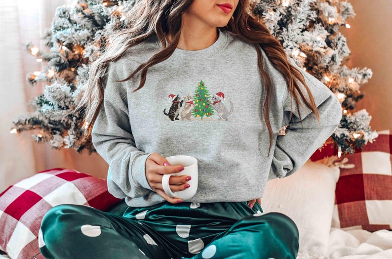 K&ouml;nnte beinhalten: Ein graues Sweatshirt mit einem gestickten Design von drei Katzen mit Weihnachtsm&uuml;tzen um einen Weihnachtsbaum. Die Katzen stehen vor einem geschm&uuml;ckten Weihnachtsbaum. Die Person h&auml;lt eine wei&szlig;e Tasse.