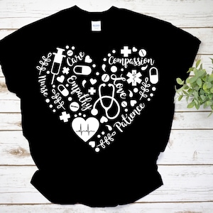 Puede incluir: Camiseta negra con un dise&ntilde;o de coraz&oacute;n blanco que presenta s&iacute;mbolos m&eacute;dicos y las palabras "Enfermera", "Cuidado", "Empat&iacute;a", "Compasi&oacute;n", "Amor" y "Paciencia".