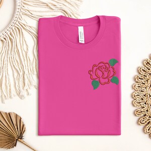 May include: A pink t-shirt with a red and green embroidered rose on the left chest.