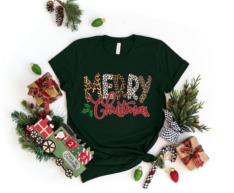 May include: A dark green t-shirt with the words "Merry Christmas" in red and white letters with a leopard print pattern. The letters are layered on top of each other and the word "Christmas" is slightly smaller than the word "Merry".