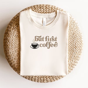 May include: A white t-shirt with the text "but first coffee" in brown lettering. A small black coffee cup with steam is above the word "coffee".