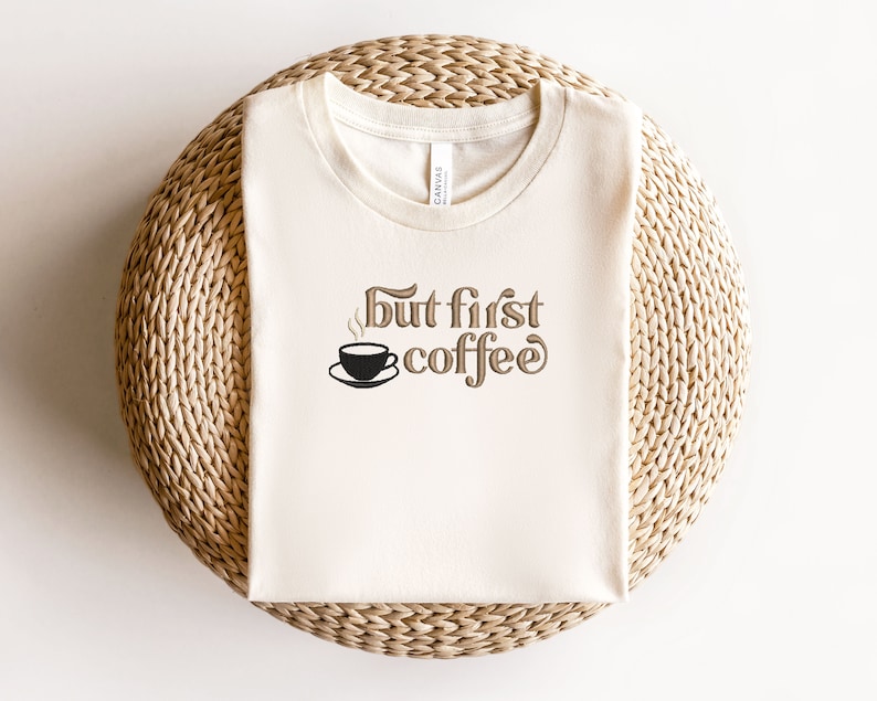 May include: A white t-shirt with the text "but first coffee" in brown lettering. A small black coffee cup with steam is above the word "coffee".
