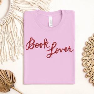 K&ouml;nnte beinhalten: Ein pinkfarbenes T-Shirt mit dem Schriftzug "Book Lover" in roter Stickerei.