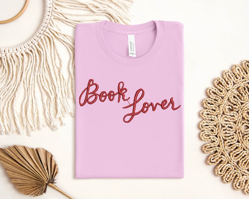 K&ouml;nnte beinhalten: Ein pinkfarbenes T-Shirt mit dem Schriftzug "Book Lover" in roter Stickerei.
