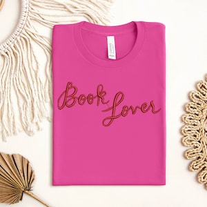 K&ouml;nnte beinhalten: Pinkes T-Shirt mit dem Schriftzug "Book Lover" in roter Stickerei.