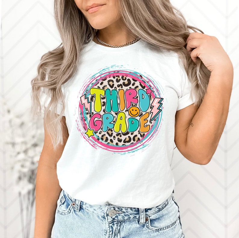 May include: White t-shirt with a colorful graphic design that says "Third Grade" in a retro style. The graphic features a leopard print background, stars, and lightning bolts.