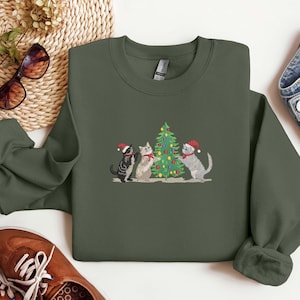 K&ouml;nnte beinhalten: Ein olivgr&uuml;nes Sweatshirt mit einem gestickten Design von drei Katzen, die einen Weihnachtsbaum schm&uuml;cken. Die Katzen tragen Weihnachtsm&uuml;tzen und Schals. Das Sweatshirt wird mit Sonnenbrille, Schuhen und Jeans pr&auml;sentiert.