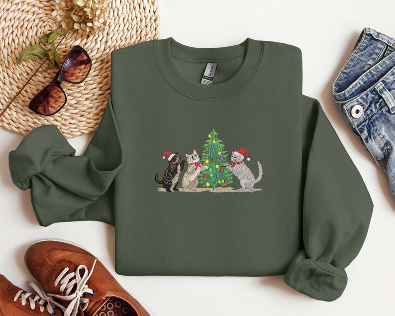 K&ouml;nnte beinhalten: Ein olivgr&uuml;nes Sweatshirt mit einem gestickten Design von drei Katzen, die einen Weihnachtsbaum schm&uuml;cken. Die Katzen tragen Weihnachtsm&uuml;tzen und Schals. Das Sweatshirt wird mit Sonnenbrille, Schuhen und Jeans pr&auml;sentiert.