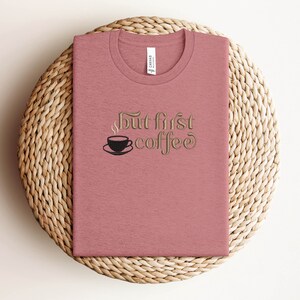 May include: A pink t-shirt with the text "But first coffee" in a gold font. A small black coffee cup with steam is above the word "coffee".