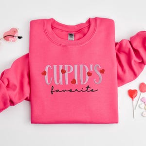 May include: Pink sweatshirt with the text "CUPID'S favorite" in black and white lettering with small red hearts around the text.