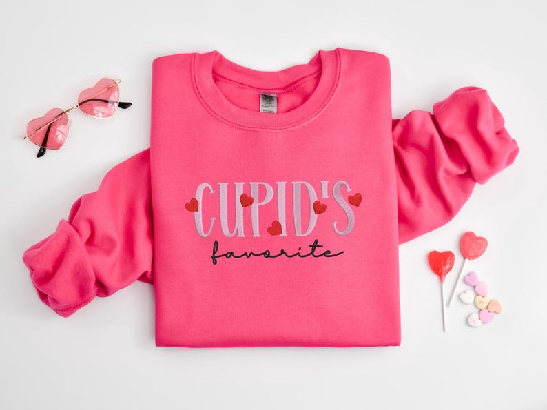 May include: Pink sweatshirt with the text "CUPID'S favorite" in black and white lettering with small red hearts around the text.