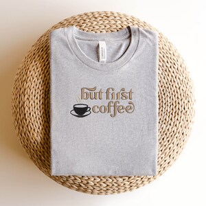 May include: A gray t-shirt with the text "But first, coffee" in brown lettering. A small black coffee cup with steam is above the text.