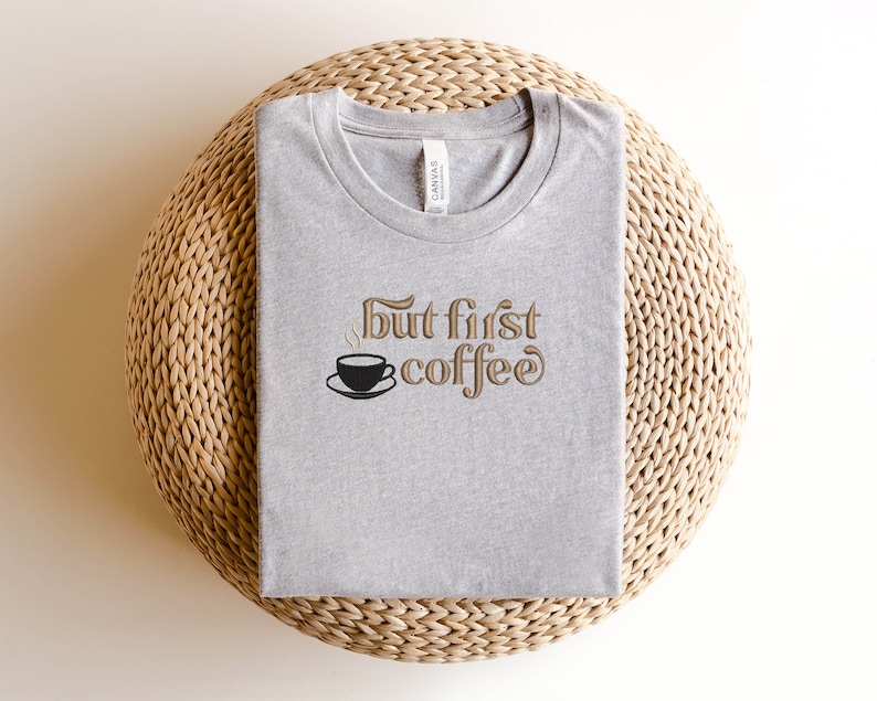 May include: A gray t-shirt with the text "But first, coffee" in brown lettering. A small black coffee cup with steam is above the text.