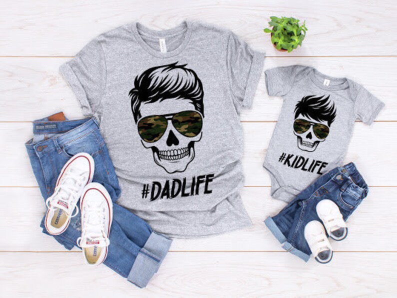 May include: Two gray t-shirts with a skull graphic and the text "#DADLIFE" and "#KIDLIFE". The shirts are laid out on a white wooden surface with blue jeans and white sneakers.