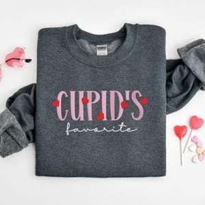 May include: A dark gray sweatshirt with the text "CUPID'S favorite" in pink lettering with red hearts.