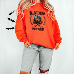 Może przedstawiać: Pomarańczowa bluza z czarnym nadrukiem graficznym przedstawiającym dynię z tekstem "Halloweentown Est. 1998 Where being normal is vastly overrated".