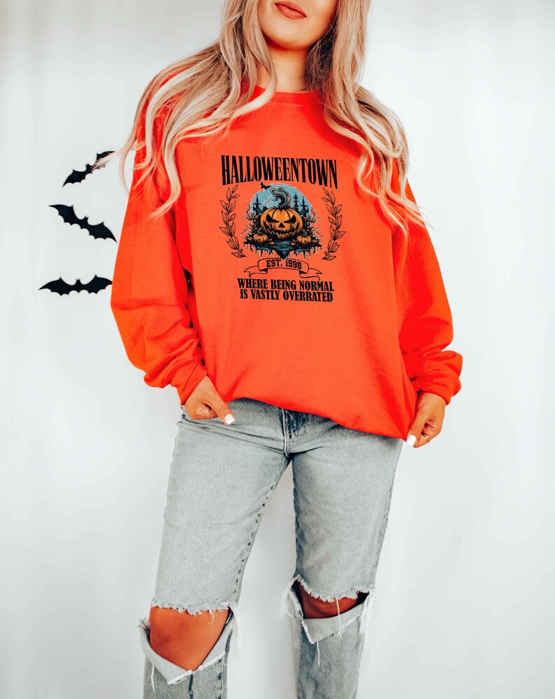 Może przedstawiać: Pomarańczowa bluza z czarnym nadrukiem graficznym przedstawiającym dynię z tekstem "Halloweentown Est. 1998 Where being normal is vastly overrated".