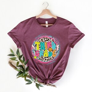 May include: A maroon t-shirt with a circle design featuring the words "Third Grade" in colorful letters with a leopard print background.
