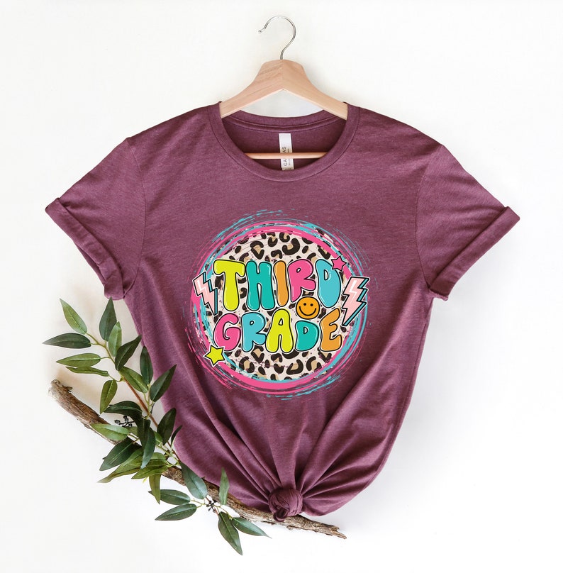 May include: A maroon t-shirt with a circle design featuring the words "Third Grade" in colorful letters with a leopard print background.
