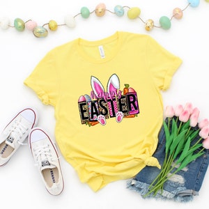 May include: Yellow t-shirt with a graphic design of bunny ears and the text "Happy Easter" in pink and white letters. The graphic also includes a carrot, eggs, and paw prints.