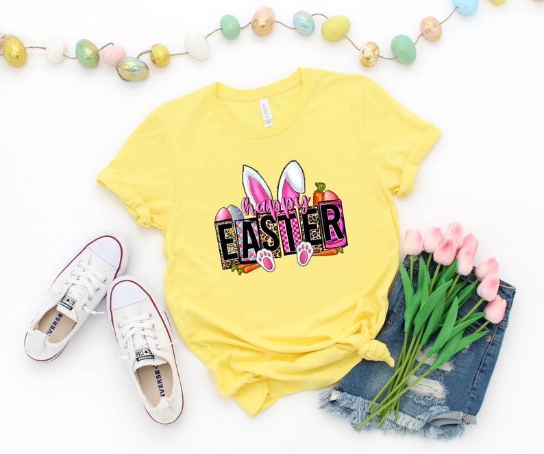May include: Yellow t-shirt with a graphic design of bunny ears and the text "Happy Easter" in pink and white letters. The graphic also includes a carrot, eggs, and paw prints.