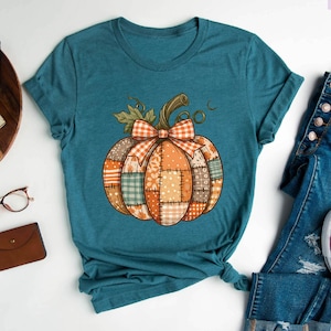 May include: Teal t-shirt featuring a patchwork pumpkin design with a bow. The pumpkin has orange, green, and tan patches. The shirt is paired with blue jeans and white sneakers. The text "Purple Rush Clothing" is visible.