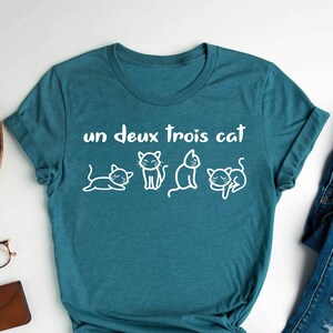 May include: Teal blue t-shirt with white text that says "un deux trois cat" and a row of four white cartoon cats.