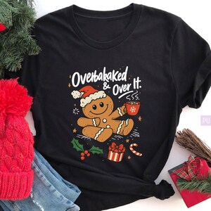 May include: Black t-shirt with a Christmas-themed graphic. The design features a gingerbread man wearing a Santa hat, holding a mug, and the text "Overbaked & Over It." The shirt is paired with a red knit hat and blue jeans.