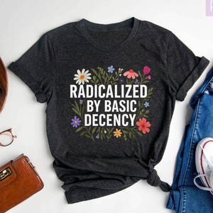 May include: Dark gray heather t-shirt with the text "RADICALIZED BY BASIC DECENCY" surrounded by colorful flowers. The shirt is styled with blue jeans and white sneakers. A brown wallet and glasses are also visible.