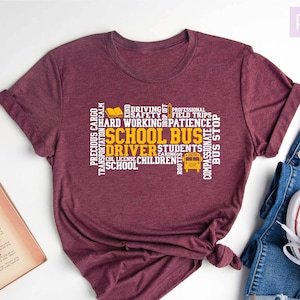 May include: A maroon t-shirt with a white graphic design that says "School Bus Driver" in a word cloud. The words in the cloud are related to the job of a school bus driver, such as "safety", "patience", "children", and "transportation".