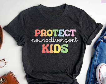 Op de afbeelding: Donkergrijs heather t-shirt met de tekst "PROTECT neurodivergent KIDS" in regenboogkleuren. Het shirt ligt plat op een wit oppervlak met een spijkerbroek, een portemonnee, een camera en een potplant.