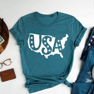 May include: A teal blue t-shirt with a white outline of the United States and the word "USA" inside the outline. The letters are decorated with white stars. The t-shirt is laid flat on a white surface with a pair of glasses, a brown wallet, a wooden tray with a camera and a watch, and a pair of blue jeans and white sneakers.