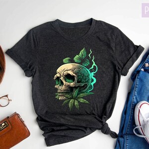 May include: Dark gray heather V-neck t-shirt with a graphic of a skull with green leaves and smoke. The shirt is laid flat on a white surface with a pair of glasses, a small brown purse, a potted plant, a vintage camera, and a pair of blue jeans with white sneakers.