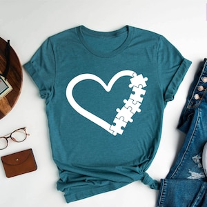 May include: Heather deep teal t-shirt with a white heart design and puzzle pieces. The shirt is paired with blue jeans and white sneakers. The words "Purple Rush Clothing" are in the top right corner.