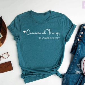 May include: Heather deep teal t-shirt with the text "Occupational Therapy is a work of heart" in white script. The shirt is styled with blue jeans and white sneakers. A wooden tray with a plant, camera, and accessories is also in the image.